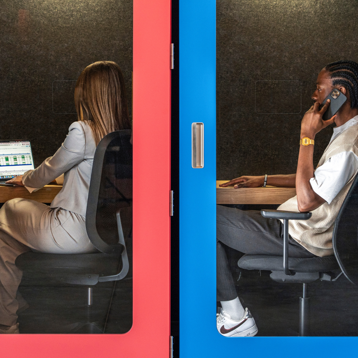 Two modern glass office pods with bold red and blue doors, shown with two people working inside on a dark textured wall background.